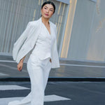 Two professional women showcasing high-end executive business suits: one in a crisp white pant suit with a shawl lapel blazers, and the other in a sophisticated camel skirt suit with a tailored vest, perfect for formal corporate settings.