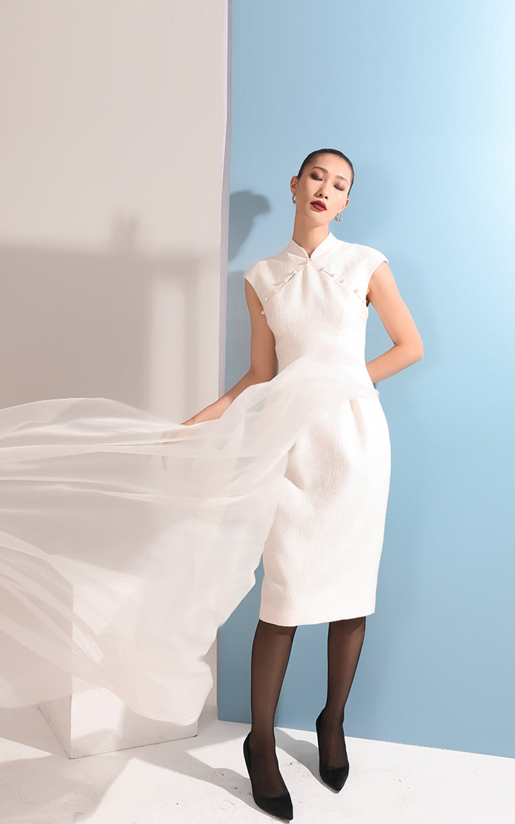 Model in white Tulip Cheongsam Dress with a tulip-shaped skirt and flowing chiffon, standing against a sky-blue backdrop, emphasizing the dress's flattering silhouette.