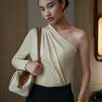 A stylish Asian woman modeling a sophisticated cream-colored asymmetric one-shoulder top featuring a single long sleeve, elegant diagonal draped pleats, and styled with high-waisted black trousers.