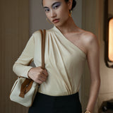 A stylish Asian woman modeling a sophisticated cream-colored asymmetric one-shoulder top featuring a single long sleeve, elegant diagonal draped pleats, and styled with high-waisted black trousers.