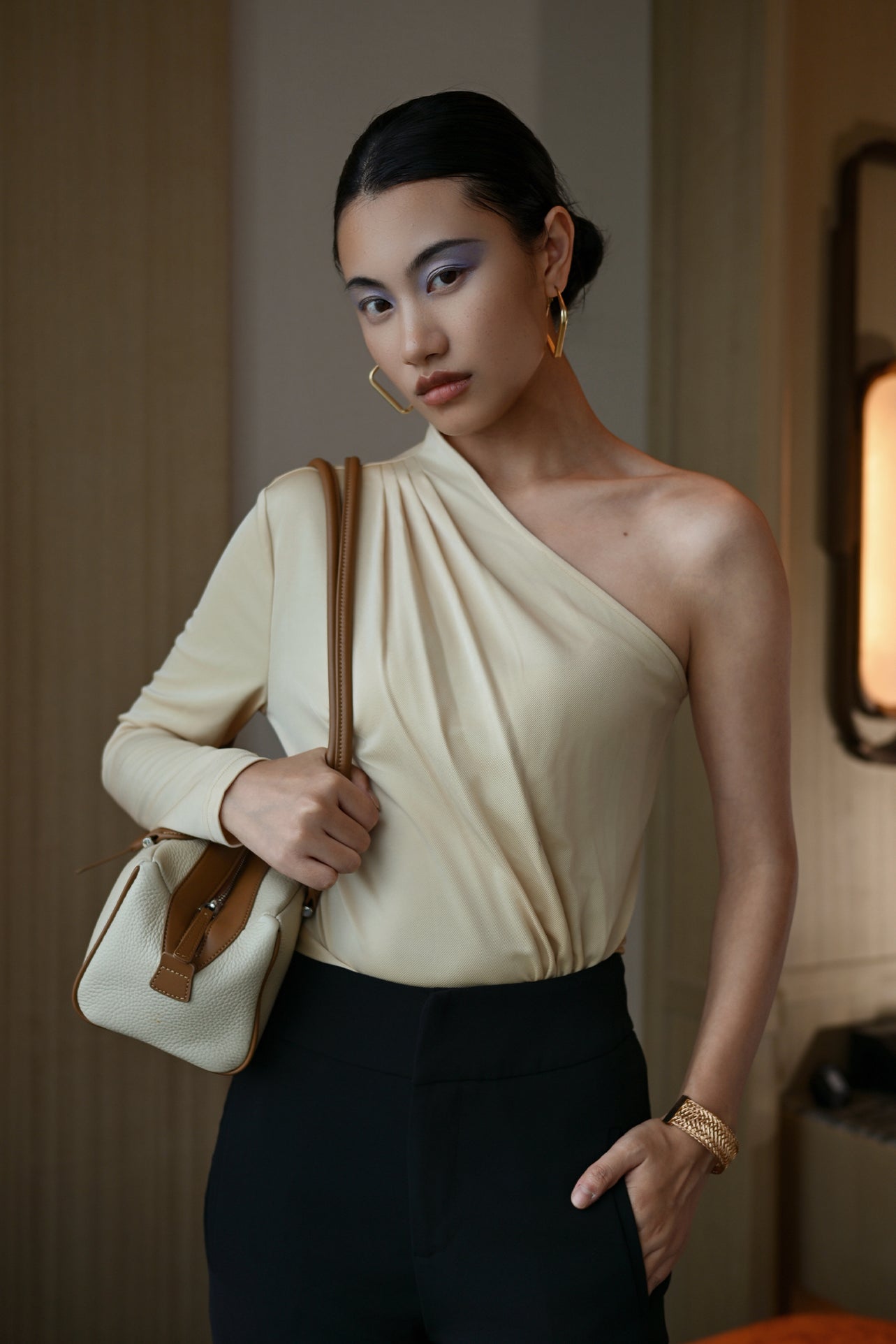 A stylish Asian woman modeling a sophisticated cream-colored asymmetric one-shoulder top featuring a single long sleeve, elegant diagonal draped pleats, and styled with high-waisted black trousers.