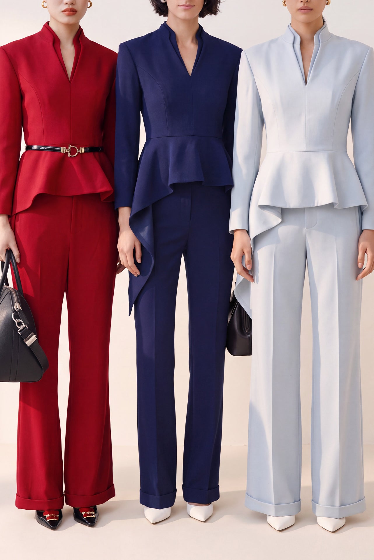 A style comparison of three women wearing tailored two-piece power suits featuring long-sleeve split V-neck jackets with dramatic asymmetrical peplum hems and matching cuffed wide-leg trousers; shown in vibrant crimson red, deep navy blue, and pale ice blue.