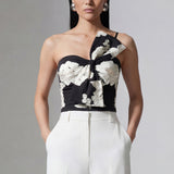 A professional woman wearing a sophisticated one-shoulder black and white floral print top, featuring a dramatic oversized bow detail on the shoulder, paired with white high-waisted trousers for an elegant evening look.