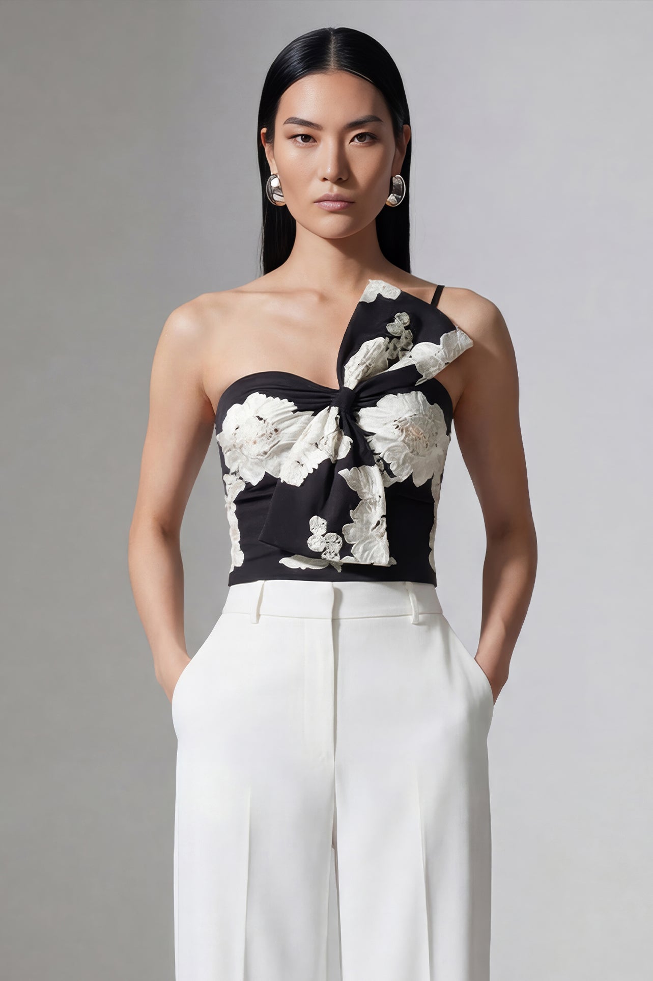 A professional woman wearing a sophisticated one-shoulder black and white floral print top, featuring a dramatic oversized bow detail on the shoulder, paired with white high-waisted trousers for an elegant evening look.