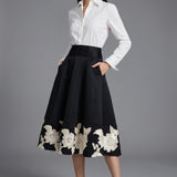 An elegant woman wearing a sophisticated black A-line midi dress, featuring exquisite white floral embroidery along the hem and professional three-quarter sleeves for a timeless formal look.