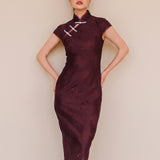 An elegant woman wearing a sophisticated burgundy jacquard modern qipao cheongsam dress featuring a classic mandarin collar, delicate contrast frog closures, cap sleeves, and a tailored sheath silhouette.