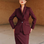 A professional woman wearing glasses and an elegant deep burgundy skirt suit featuring a sculptural stand-up lapel blazer with a single-button closure, tailored flap pockets, and a matching high-waisted pencil skirt.