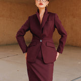 A professional woman wearing glasses and an elegant deep burgundy skirt suit featuring a sculptural stand-up lapel blazer with a single-button closure, tailored flap pockets, and a matching high-waisted pencil skirt.