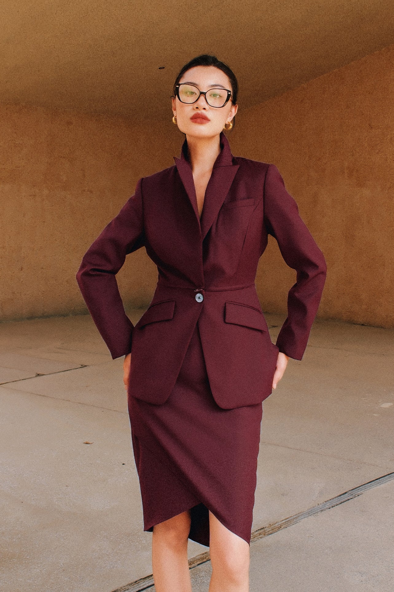 A professional woman wearing glasses and an elegant deep burgundy skirt suit featuring a sculptural stand-up lapel blazer with a single-button closure, tailored flap pockets, and a matching high-waisted pencil skirt.
