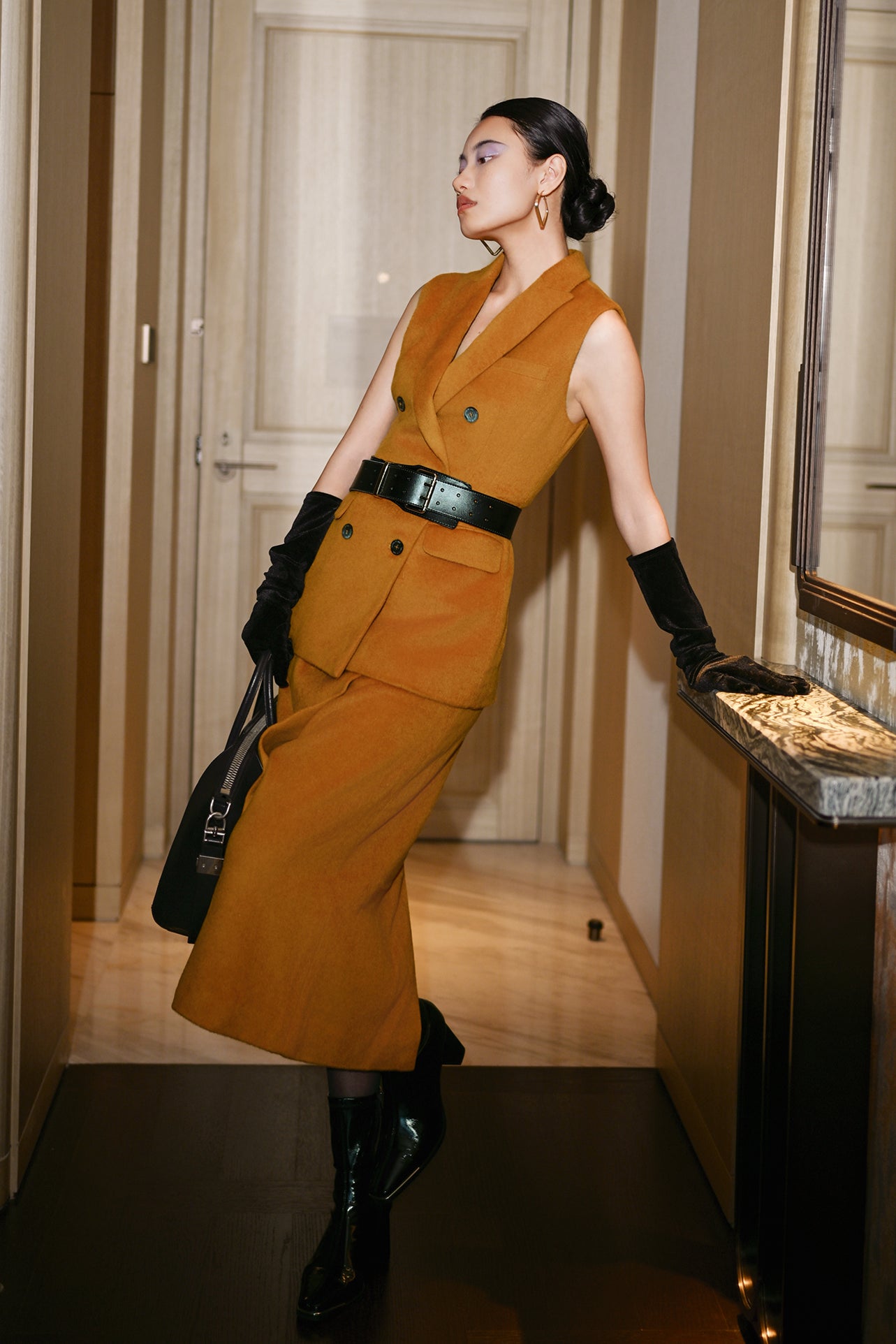 An elegant Asian woman posing in a luxurious camel wool two-piece suit featuring a sleeveless double-breasted longline vest cinched with a wide black belt, paired with a matching midi column skirt and black opera gloves.