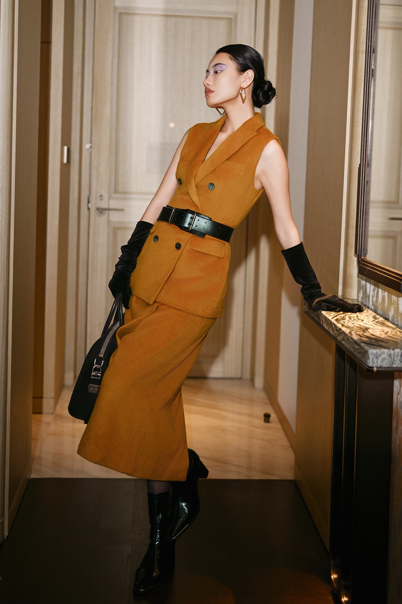 An elegant Asian woman posing in a luxurious camel wool two-piece suit featuring a sleeveless double-breasted longline vest cinched with a wide black belt, paired with a matching midi column skirt and black opera gloves.