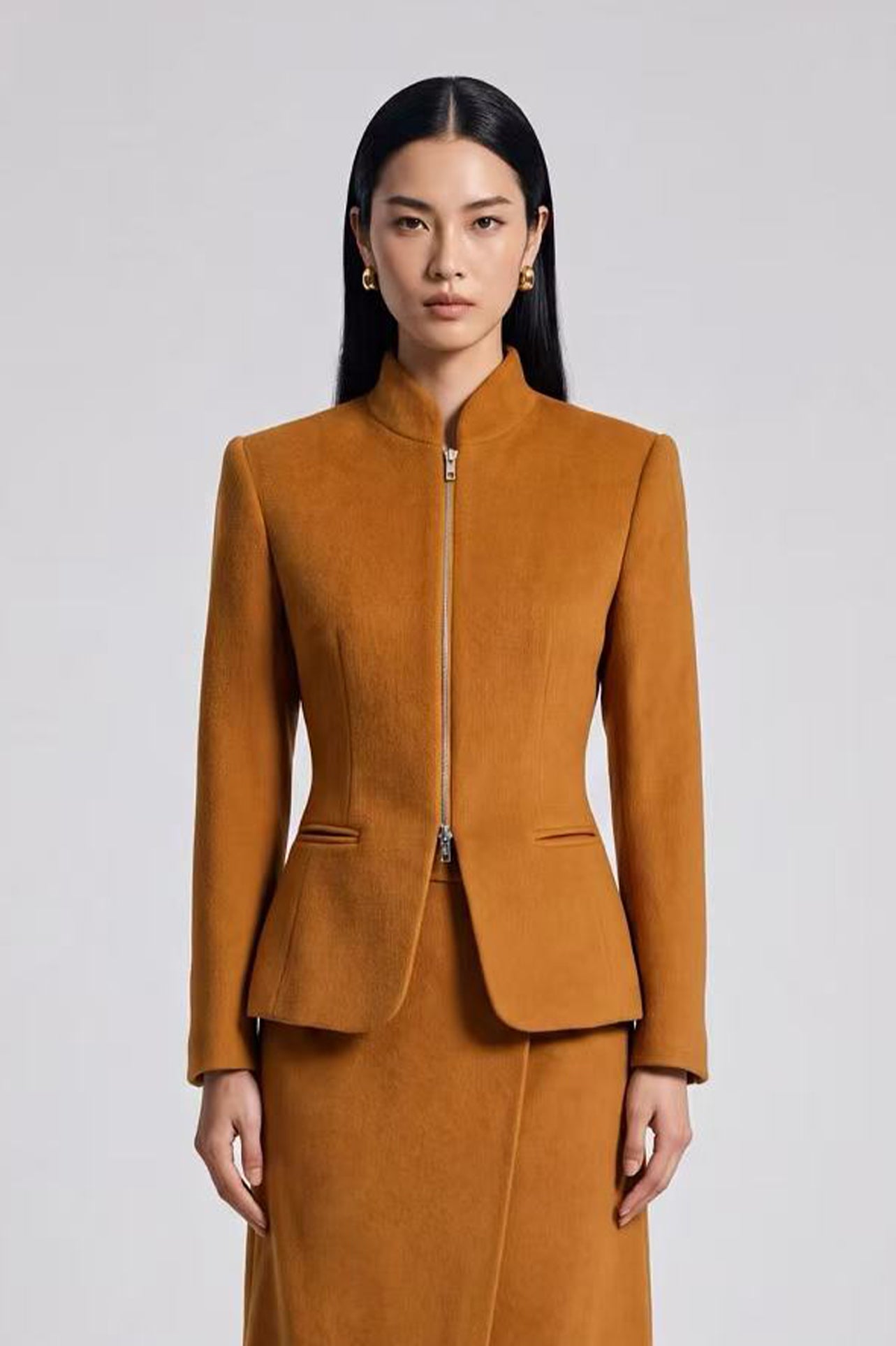 A chic professional Asian woman wearing a luxurious camel wool tailored jacket featuring a sleek stand collar, exposed two-way front zipper, structured shoulders, and an hourglass silhouette paired with a matching skirt.