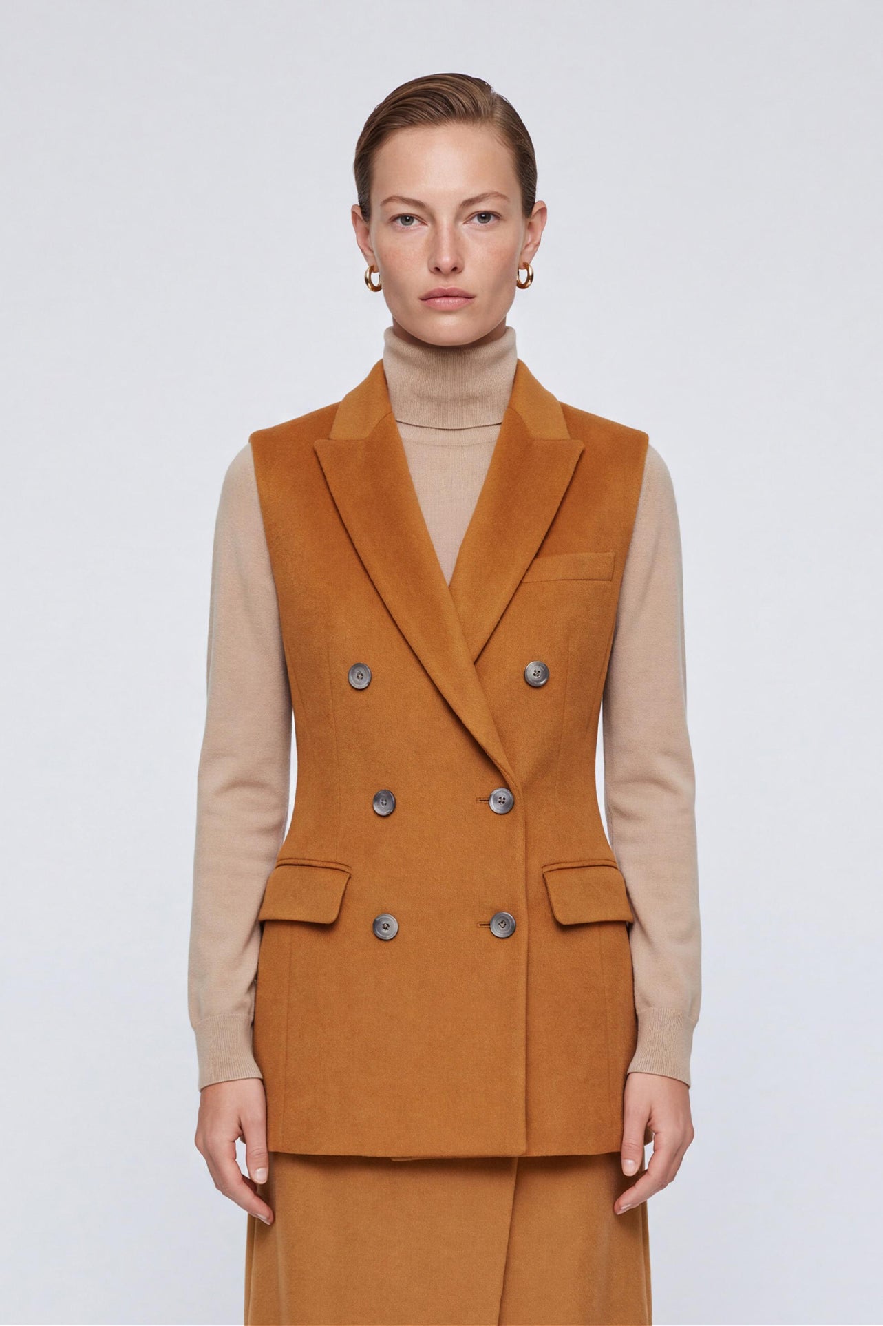 A chic professional woman wearing a tailored camel double-breasted longline suit vest featuring classic notch lapels and flap pockets, layered over a soft beige turtleneck sweater for a sophisticated quiet luxury look.