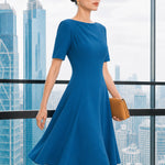 A beautiful model wearing a sophisticated medium blue A-line midi dress featuring an elegant boat neckline with refined asymmetric shoulder pleating, short sleeves, and a flowing flared swing skirt.