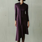 An elegant woman wearing a sophisticated deep plum-purple long-sleeve knit midi dress featuring a draped high-neck tie scarf and a dramatic asymmetrical flounce mermaid hemline.
