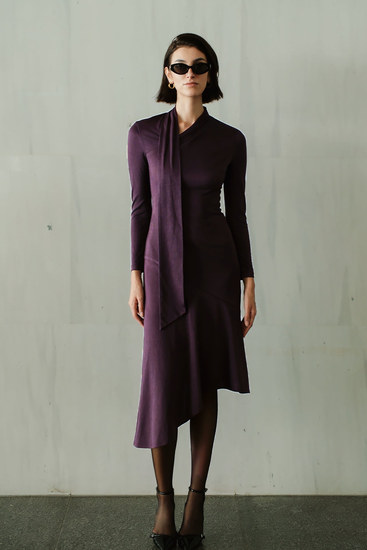 An elegant woman wearing a sophisticated deep plum-purple long-sleeve knit midi dress featuring a draped high-neck tie scarf and a dramatic asymmetrical flounce mermaid hemline.