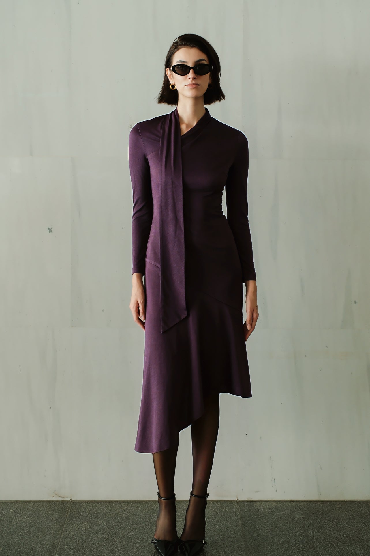 An elegant woman wearing a sophisticated deep plum-purple long-sleeve knit midi dress featuring a draped high-neck tie scarf and a dramatic asymmetrical flounce mermaid hemline.