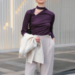 A chic woman wearing an elegant deep plum long-sleeve blouse with an asymmetric high-neck scarf tie, neatly tucked into a high-waisted black tweed skirt embellished with subtle sparkling sequins.