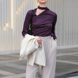 A chic woman wearing an elegant deep plum long-sleeve blouse with an asymmetric high-neck scarf tie, neatly tucked into a high-waisted black tweed skirt embellished with subtle sparkling sequins.