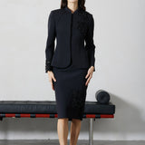 A professional woman wearing a sophisticated black skirt suit, featuring a mandarin collar with elegant leaf embroidery on the shoulder and skirt, and traditional frog button detailing for a modern oriental executive look.