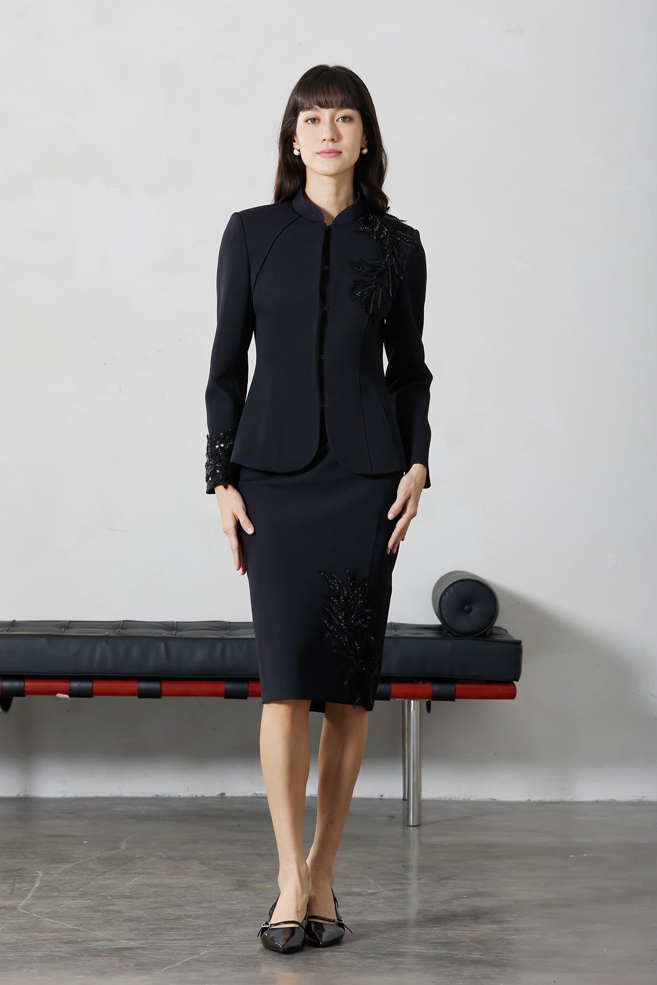 A professional woman wearing a sophisticated black skirt suit, featuring a mandarin collar with elegant leaf embroidery on the shoulder and skirt, and traditional frog button detailing for a modern oriental executive look.