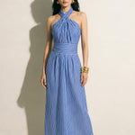 A model showcasing an elegant blue and white vertical striped maxi dress, featuring a sophisticated criss-cross halter neckline, a structured wide banded waist for a slimming silhouette, and a long flowing skirt, paired with white strap high heels.