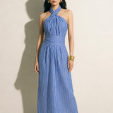 A model showcasing an elegant blue and white vertical striped maxi dress, featuring a sophisticated criss-cross halter neckline, a structured wide banded waist for a slimming silhouette, and a long flowing skirt, paired with white strap high heels.