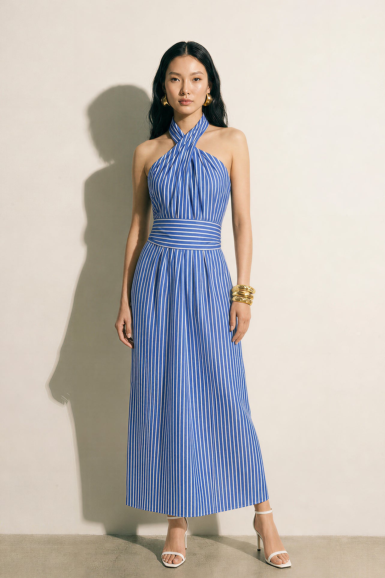 A model showcasing an elegant blue and white vertical striped maxi dress, featuring a sophisticated criss-cross halter neckline, a structured wide banded waist for a slimming silhouette, and a long flowing skirt, paired with white strap high heels.