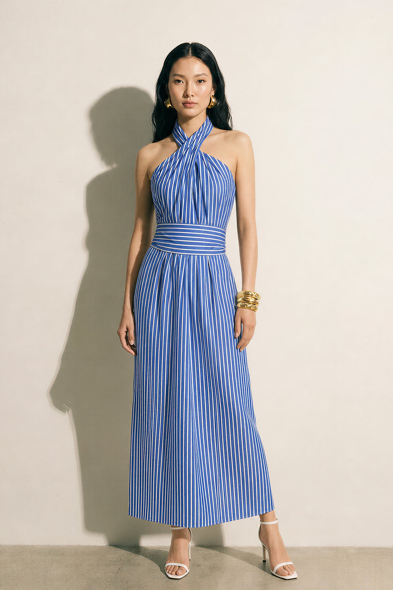 A model showcasing an elegant blue and white vertical striped maxi dress, featuring a sophisticated criss-cross halter neckline, a structured wide banded waist for a slimming silhouette, and a long flowing skirt, paired with white strap high heels.