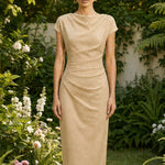 A professional woman modeling an elegant pastel pink satin midi dress, featuring a sophisticated cowl neckline, delicate cap sleeves, and slimming asymmetric ruched drapes across the waist for a quiet luxury look.