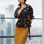 A stylish woman posing against a city skyline wearing a navy blue wrap blouse with golden ginkgo leaf print tied at the waist, paired with tailored mustard yellow wide-leg trousers.