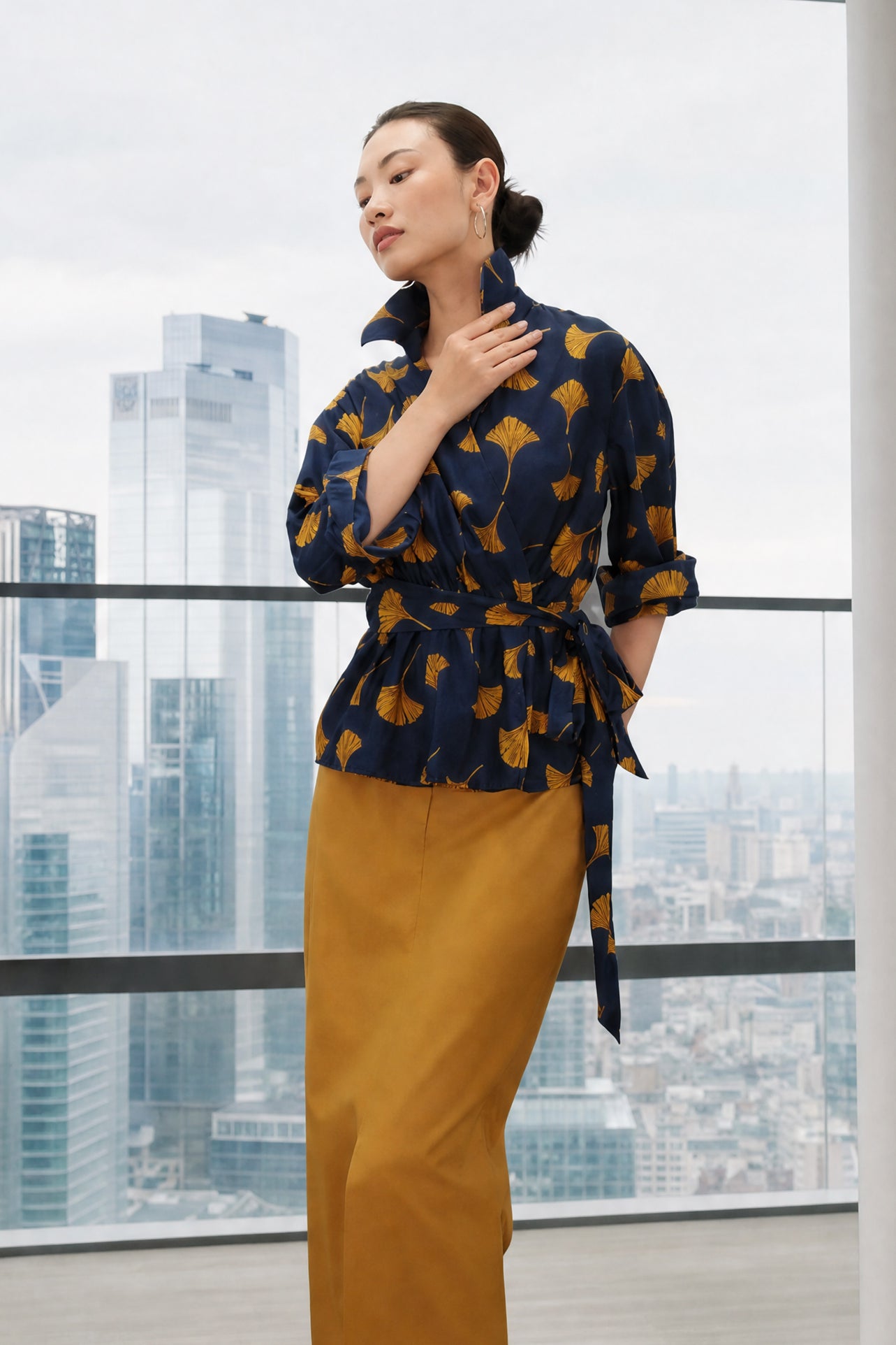A stylish woman posing against a city skyline wearing a navy blue wrap blouse with golden ginkgo leaf print tied at the waist, paired with tailored mustard yellow wide-leg trousers.