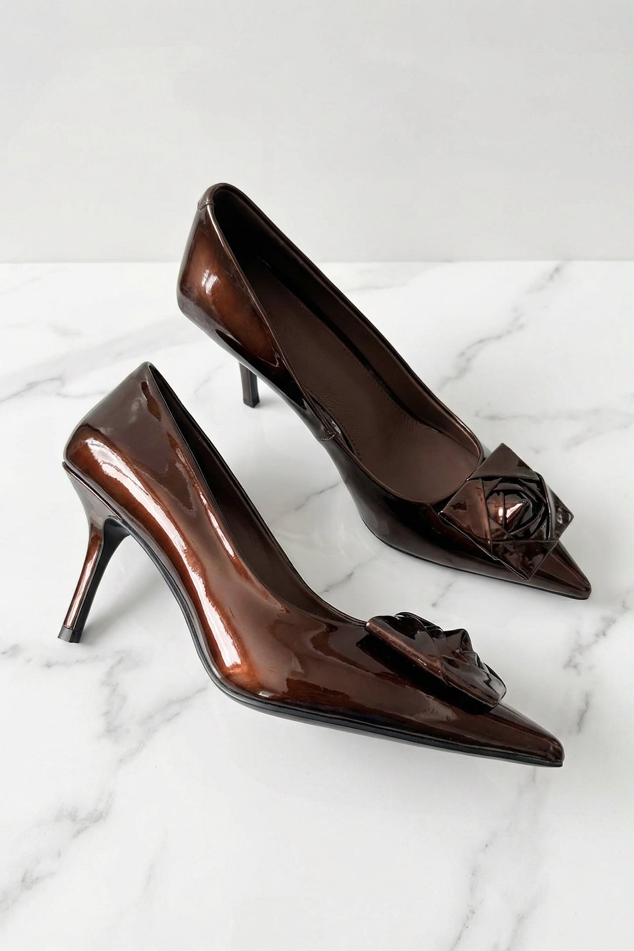 A pair of elegant dark chocolate brown patent leather pumps featuring a pointed toe and a sophisticated 3D rose floral decoration on the front, perfect for a quiet luxury office or evening look.