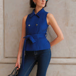 A style comparison of two professional women wearing modern cobalt blue outerwear featuring self-tie waist belts; on the left, a tailored cropped capelet jacket paired with jeans, and on the right, a double-breasted sleeveless vest with white contrast buttons styled with black trousers.