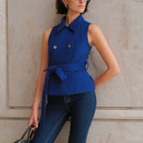 A style comparison of two professional women wearing modern cobalt blue outerwear featuring self-tie waist belts; on the left, a tailored cropped capelet jacket paired with jeans, and on the right, a double-breasted sleeveless vest with white contrast buttons styled with black trousers.