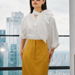 A style collection of three models presenting elegant dopamine office wear; the left in a white bow-tie blouse with a mustard yellow front-slit midi skirt, the center wearing a blue striped pussy-bow shirt with a straw hat and white wide-leg trousers, and the right in a vibrant orange satin blouse with a royal blue pencil skirt.