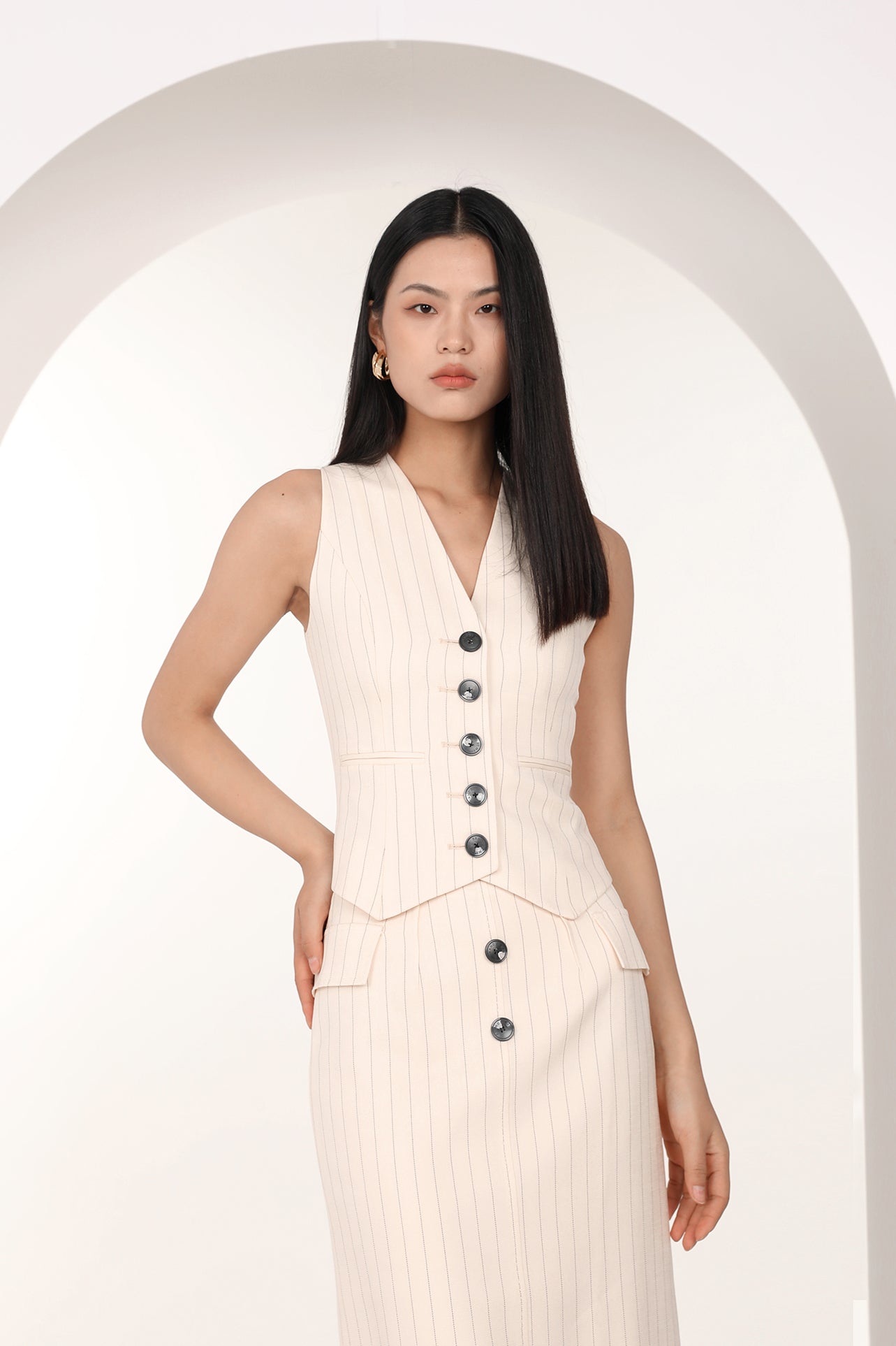 A professional woman wearing a sophisticated off-white pinstripe vest top, featuring a slim-fit tailored silhouette, classic V-neck, and contrasting black buttons for a modern business casual look.