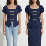 A professional woman modeling an elegant navy blue lace short top, featuring a retro square neckline, romantic short puff sleeves, and a series of pearl and rhinestone frog closures on the front with a tailored triangular split hem, paired with light blue high-waisted jeans.