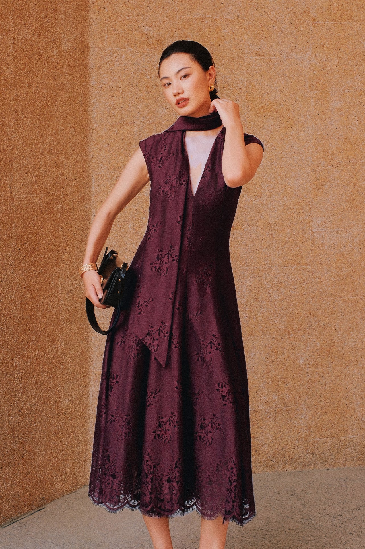 A chic woman wearing sunglasses and an elegant deep burgundy sleeveless V-neck A-line midi dress featuring textured jacquard fabric, delicate lace hemline, and a matching flowing neck scarf.