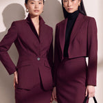 A professional woman wearing glasses and an elegant deep burgundy skirt suit featuring a sculptural stand-up lapel blazer with a single-button closure, tailored flap pockets, and a matching high-waisted pencil skirt.