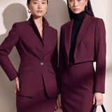 A professional woman wearing glasses and an elegant deep burgundy skirt suit featuring a sculptural stand-up lapel blazer with a single-button closure, tailored flap pockets, and a matching high-waisted pencil skirt.