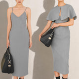 A stylish woman walking on a city street wearing an elegant light grey sheath midi dress with a sophisticated capelet sleeve design, round neckline, and side slit, carrying a black leather handbag.