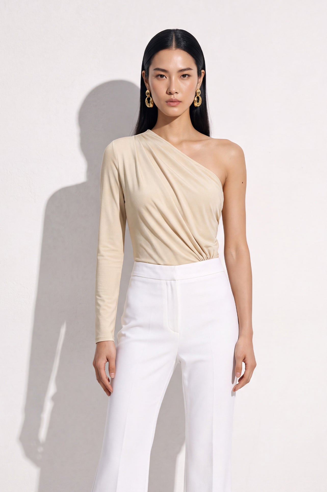 A stylish Asian woman modeling a sophisticated cream-colored asymmetric one-shoulder top featuring a single long sleeve, elegant diagonal draped pleats, and styled with high-waisted black trousers.