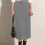 A stylish woman wearing a sophisticated grey pinstripe high-waisted midi skirt with a front slit, paired with a white button-up shirt rolled at the sleeves and black ankle boots for a minimalist corporate chic look.