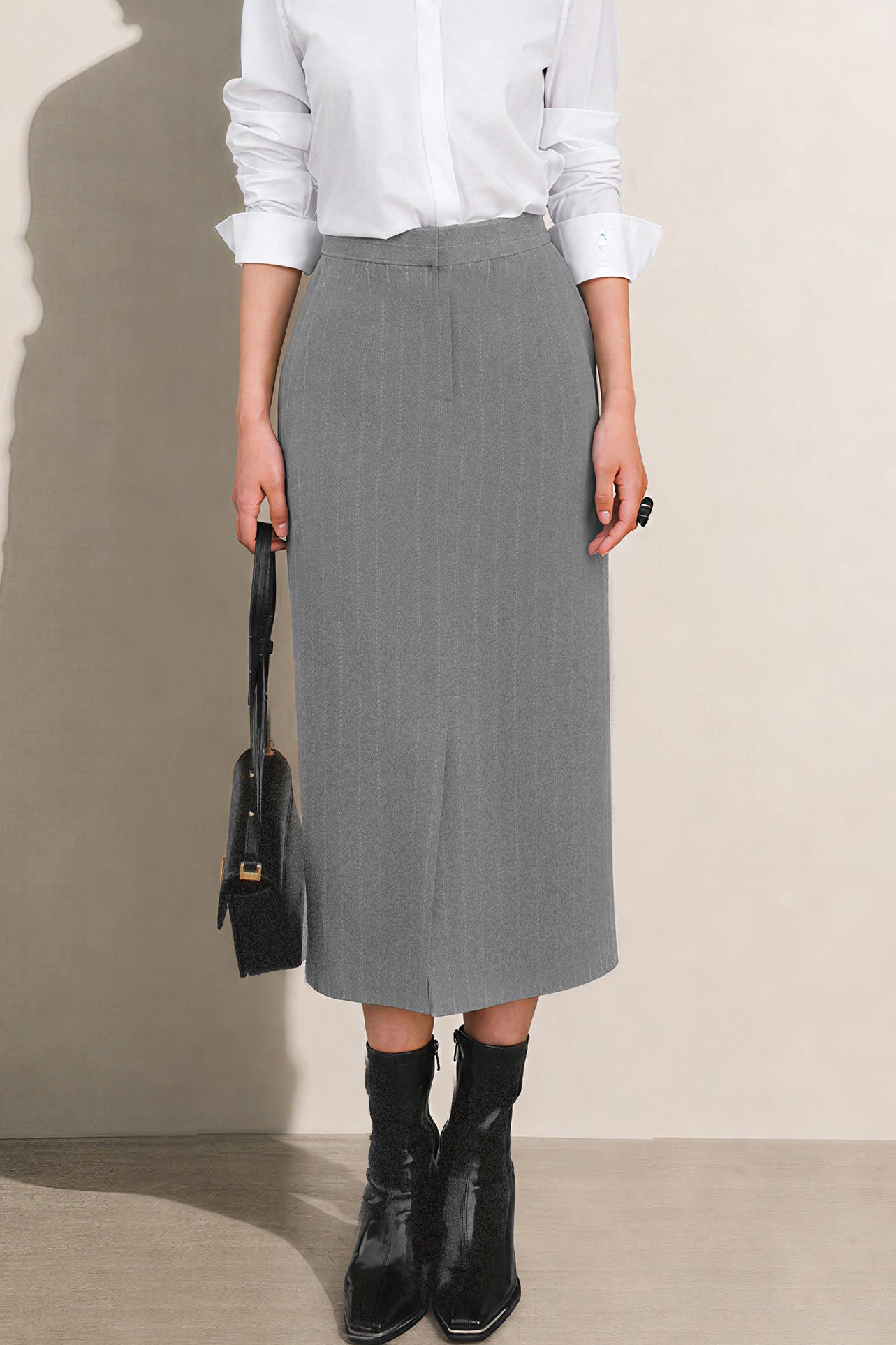 A stylish woman wearing a sophisticated grey pinstripe high-waisted midi skirt with a front slit, paired with a white button-up shirt rolled at the sleeves and black ankle boots for a minimalist corporate chic look.