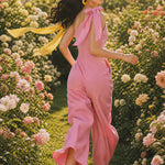 Two joyful women wearing elegant one-shoulder jumpsuits with large tie-bow shoulder straps in a sunny park; one jumps in a pastel pink wide-leg jumpsuit with yellow sneakers, while the other claps in a beige ankle-length jumpsuit.