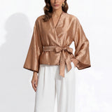 Two professional models wearing elegant shimmering satin wrap tops with kimono-style sleeves and waist ties, one in champagne gold and the other in charcoal grey with contrast white piping, paired with ivory high-waisted trousers.