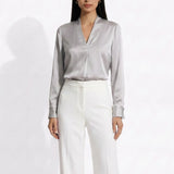 Two professional models wearing elegant V-neck silk satin blouses in shimmering silver and deep purple, featuring pleated front details and paired with white high-waisted trousers and a buttoned skirt.