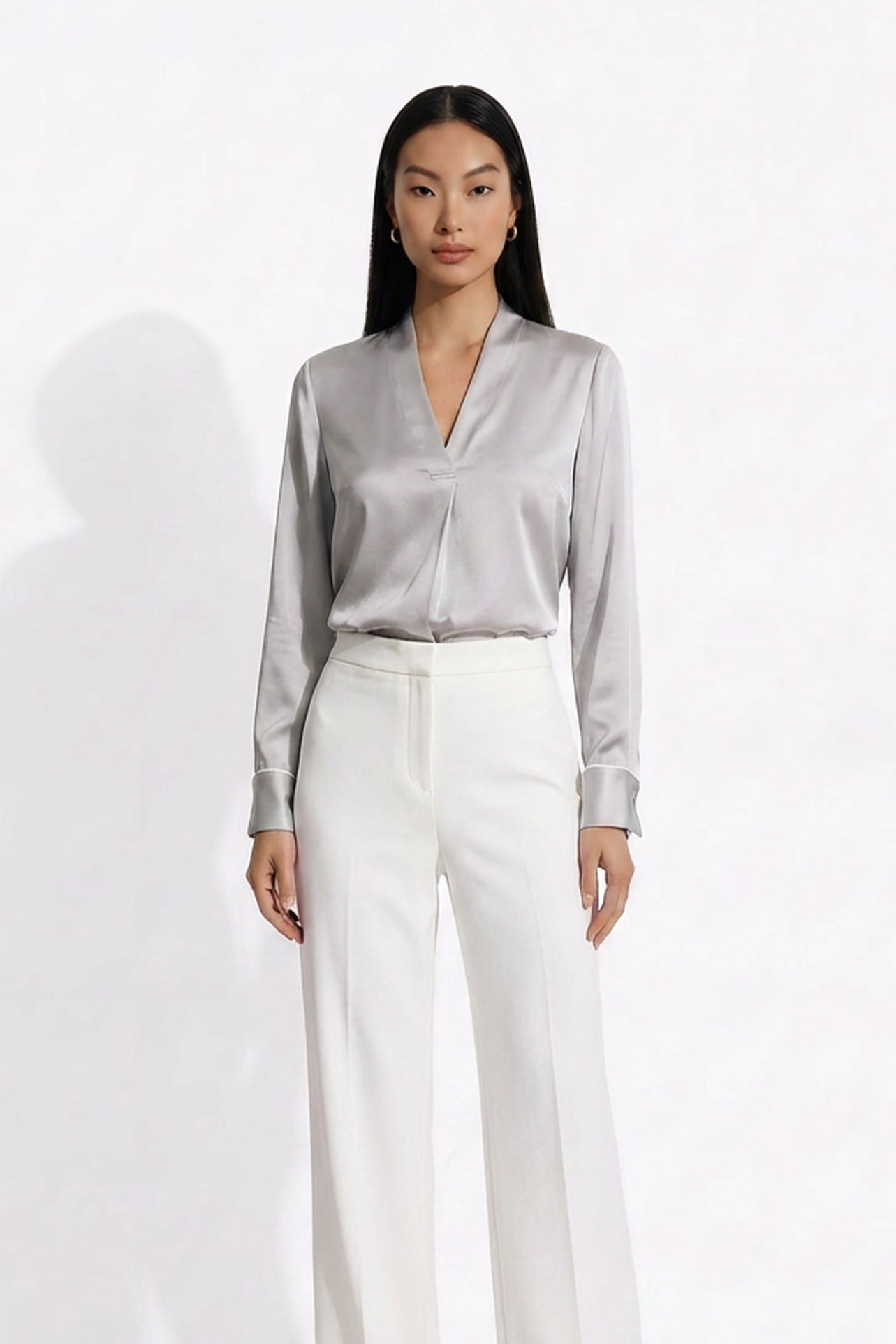 Two professional models wearing elegant V-neck silk satin blouses in shimmering silver and deep purple, featuring pleated front details and paired with white high-waisted trousers and a buttoned skirt.