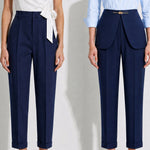 A model showcasing elegant navy blue high-waisted work trousers, featuring a unique curved overlay panel design at the waist, a slim golden metal bar buckle, pressed front creases, and chic cuffed hems, paired with a light blue button-up shirt.